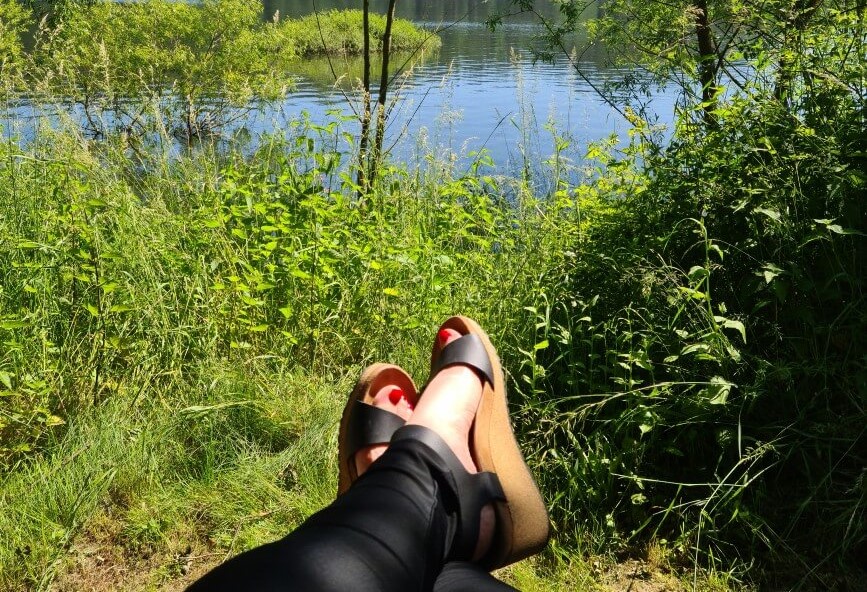Grüne Landschaft mit See im Hintergrund, Beine und Schuhe in Sandalen im Vordergrung