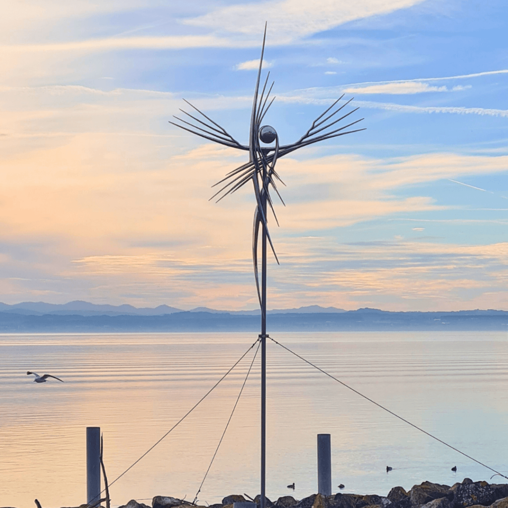 Figur vor See, im Hintergrund Gebirge und ein vorbei fliegender Vogel